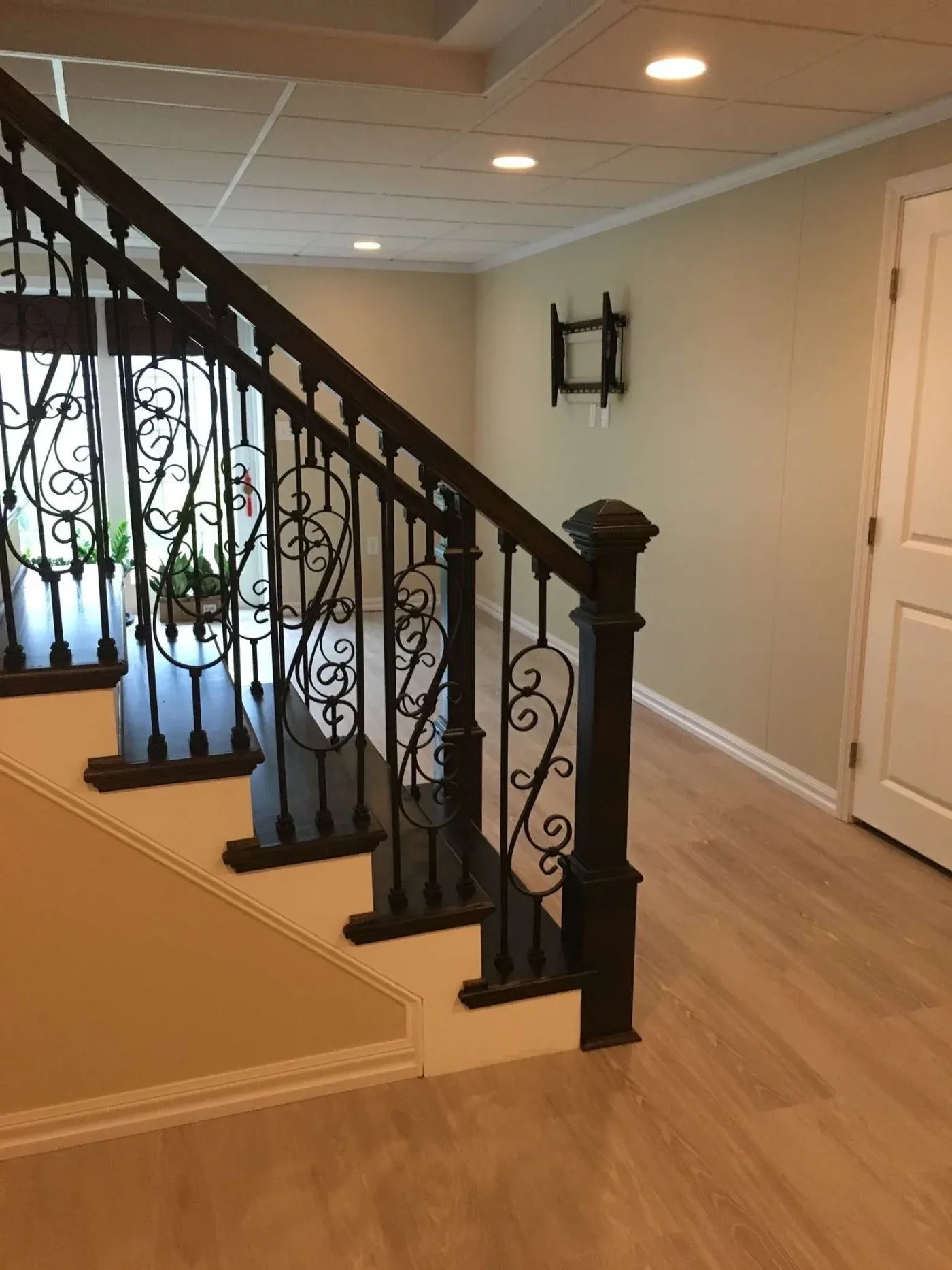 A basement with light wood flooring, featuring a staircase with ornate black metal railings. The walls are beige, creating a warm and inviting atmosphere.
