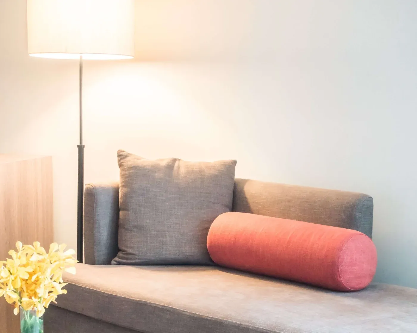 Cozy room with a gray sofa, accented by a red cylindrical pillow and matching gray square cushion. A bright floor lamp illuminates yellow flowers nearby.