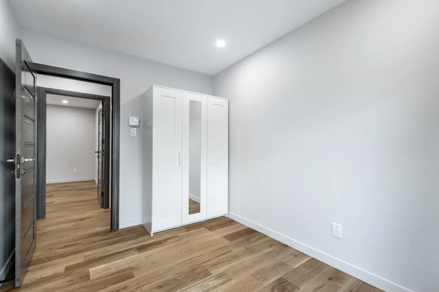 A minimalist room with light gray walls, wooden floor, and a white wardrobe with mirrors. An open dark door leads to a hallway. Bright and spacious.