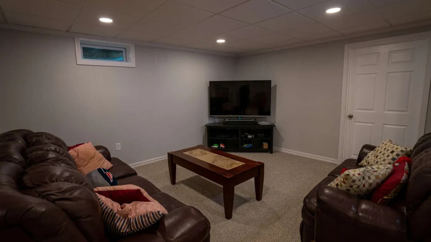 Cozy basement living room with brown leather sofas, colorful pillows, a wooden coffee table, and a flat-screen TV on a stand, under soft lighting.