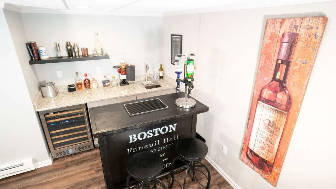 Home bar with a Boston theme, featuring a granite countertop, spirits display, and a mini fridge. Wall decor includes a vintage bottle painting.