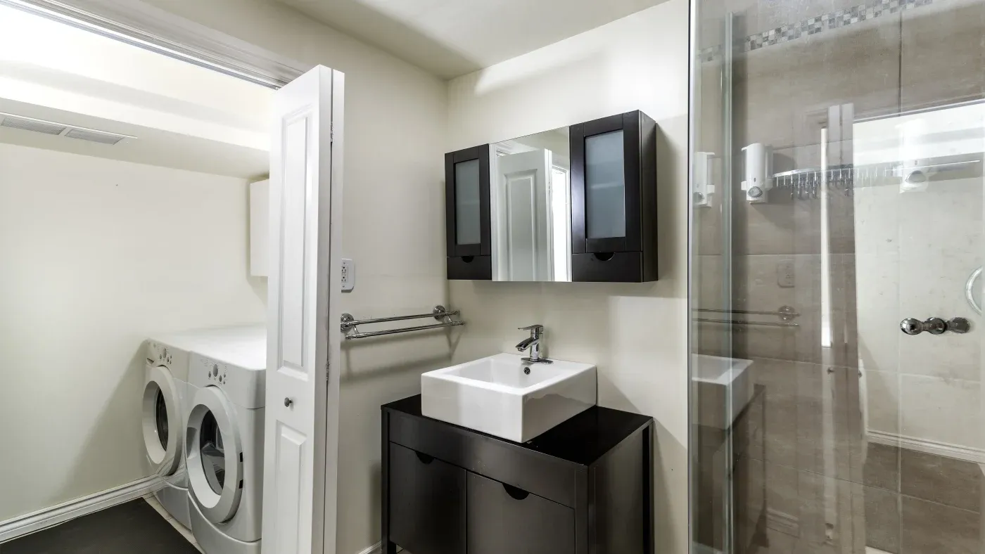 Modern bathroom with a washing machine and dryer, dark wood vanity, square sink, mirrored cabinet, and glass shower. Bright, clean, and minimalistic.
