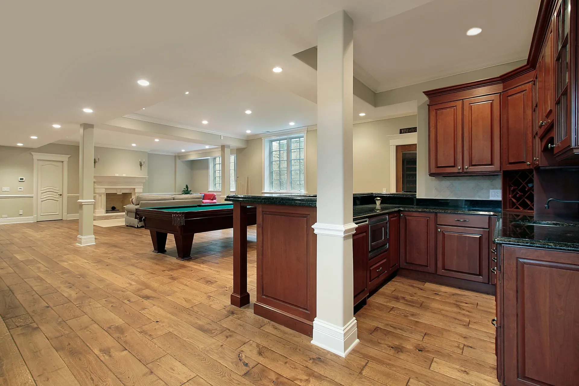 Spacious and elegant basement with wooden floors, featuring a pool table, cozy fireplace, and dark wood cabinets in a kitchenette area.