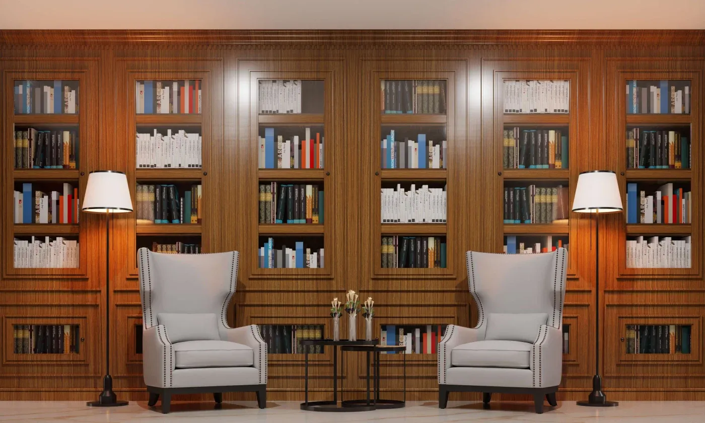 Elegant library room with wooden bookshelves filled with colorful books, two large armchairs, side tables, and a warm ambient glow from two floor lamps.