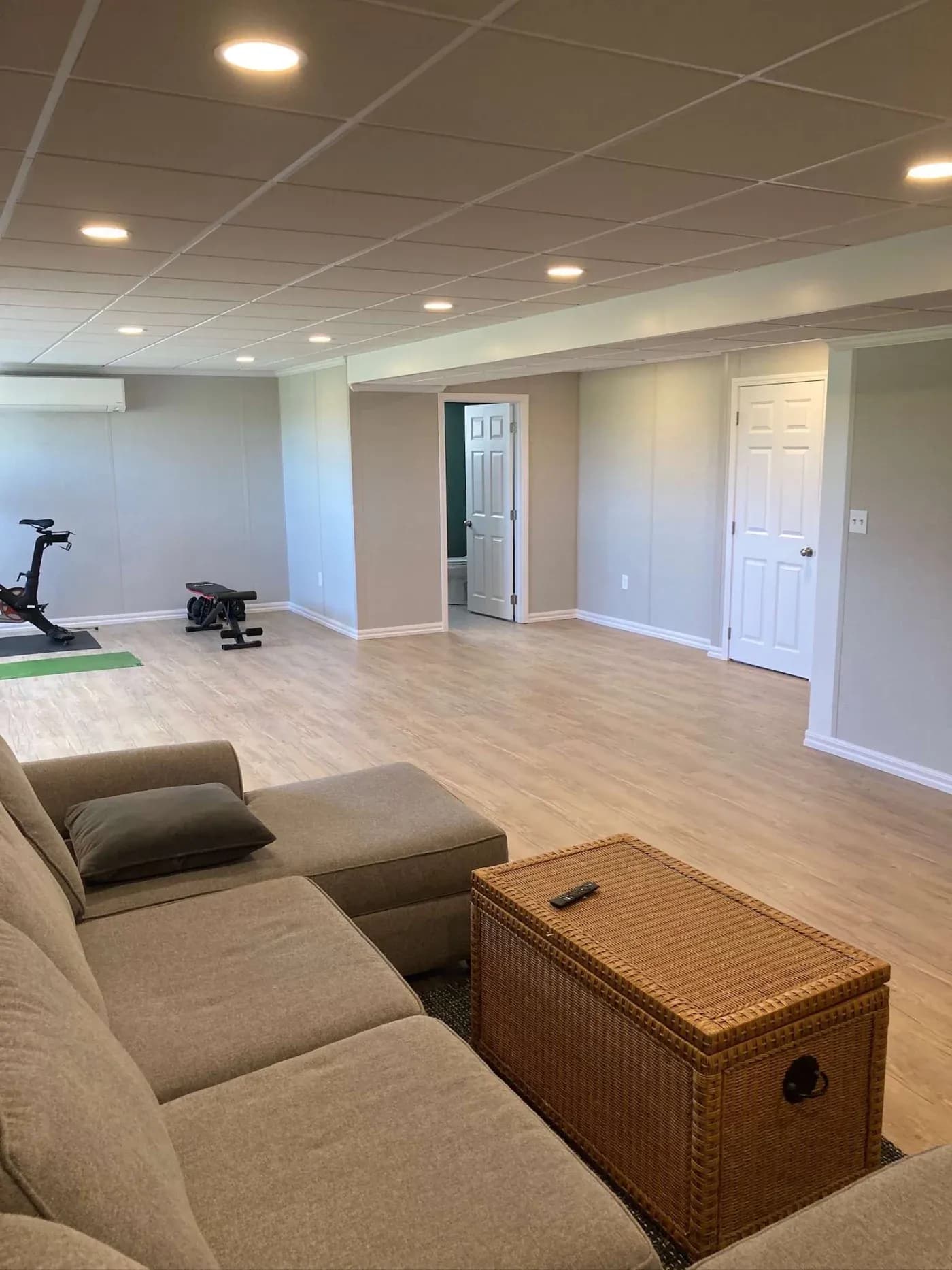 A spacious, well-lit basement with light wood flooring and neutral walls. It features a gray sectional sofa, wicker table, and exercise bike, creating a cozy and functional atmosphere.