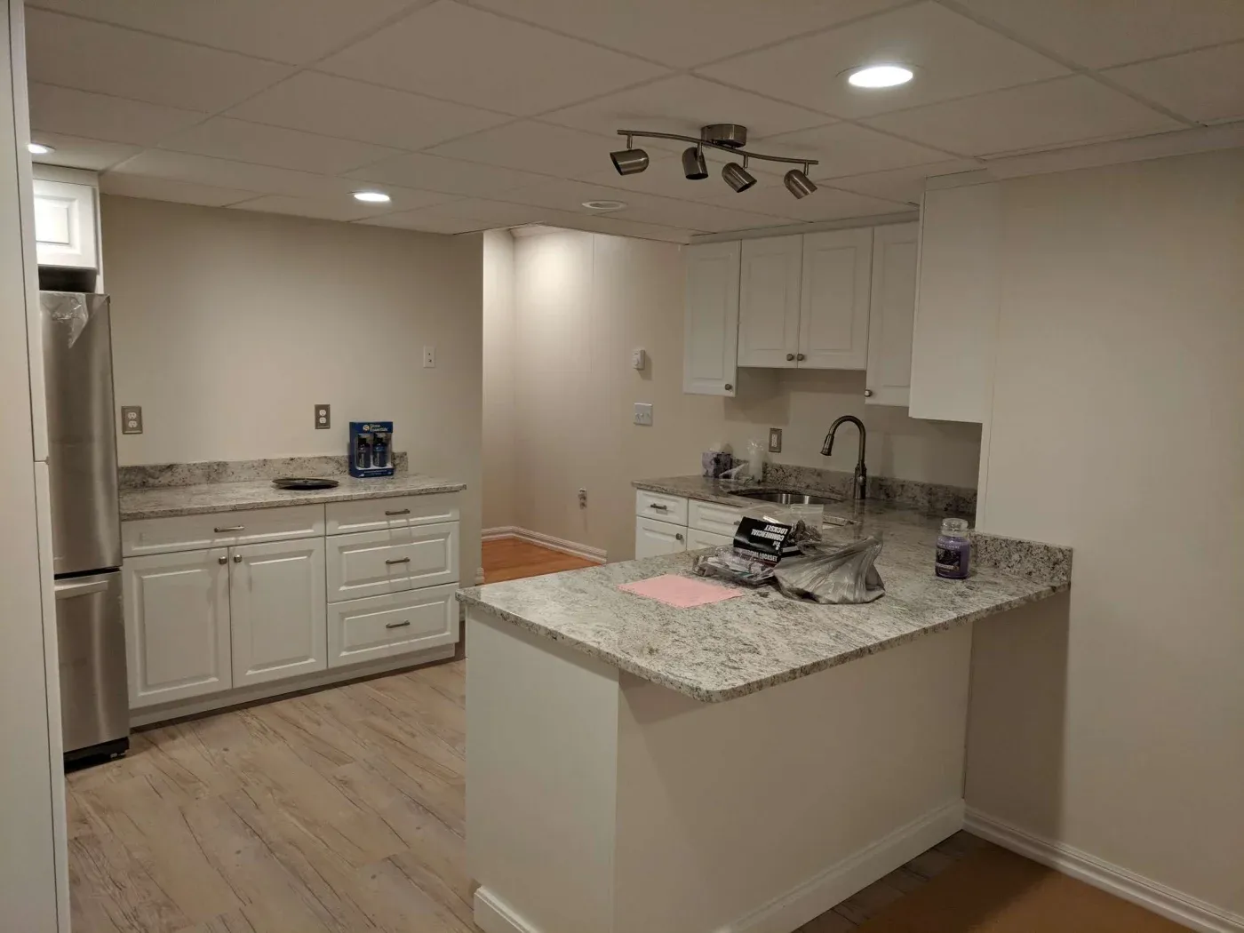 Modern kitchen with white cabinets, stainless steel fridge, and granite countertops. Wooden floor and recessed lighting create a clean, inviting space.