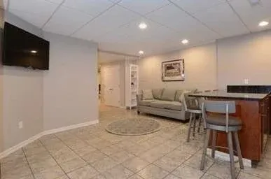 A cozy basement room with tiled floors features a wall-mounted TV, a gray sofa, a circular rug, and a small bar area with stools under soft lighting.