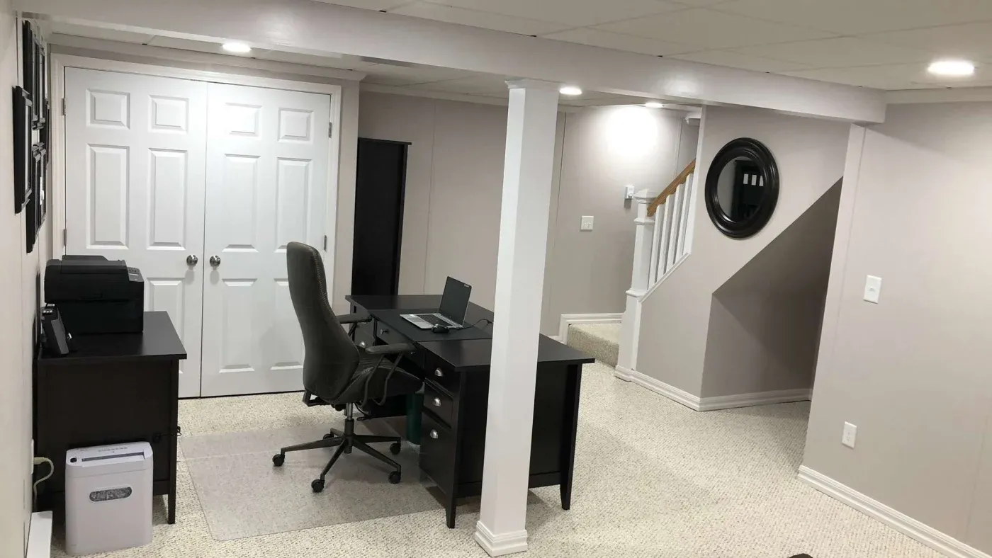 Basement home office with a laptop on a dark desk, swivel chair, and printer. White walls, carpeted floor, and a staircase with a circular mirror.