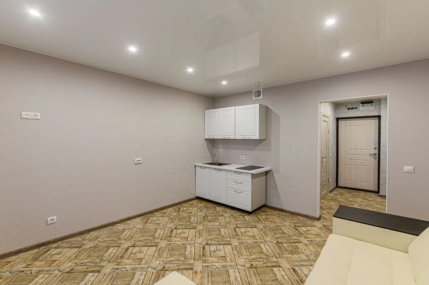 Minimalist room with beige walls and wood-patterned floor. Features small white kitchenette, ceiling lights, and cream sofa. Door leads to hallway.