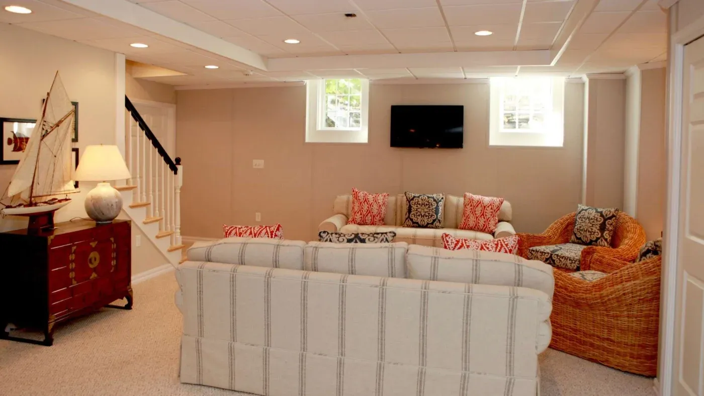 Cozy basement living room with beige walls and ceiling, light-striped sofas, and wicker chairs. Nautical decor includes a model sailboat. Natural light from small windows adds warmth.