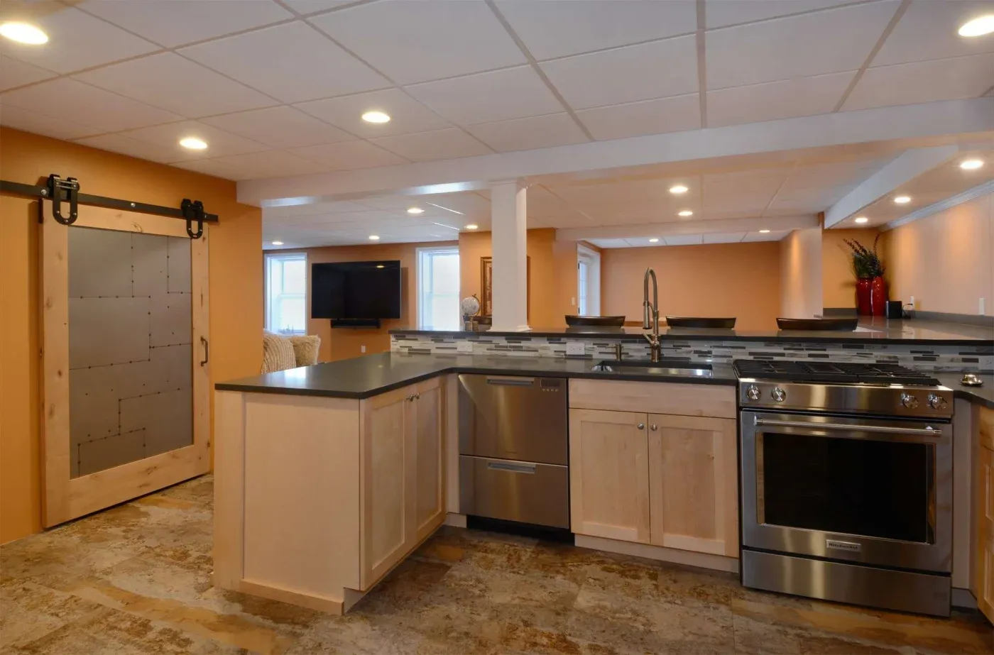 Modern basement kitchen with light wood cabinets, stainless steel appliances, and black countertops. Orange walls add warmth. Sliding barn door on left.