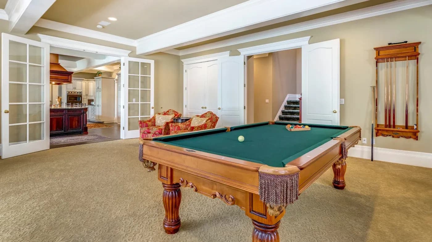 Luxurious game room with a wooden pool table in the foreground, two open French doors leading to a kitchen, plush armchairs, and a billiard cue rack on the wall.