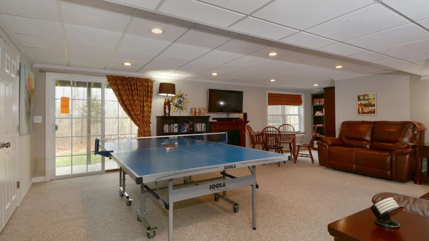 Basement game room with a blue ping pong table in the center, brown leather sofa, TV on the wall, bookshelves, wooden dining set, and sliding doors. Cozy ambiance.