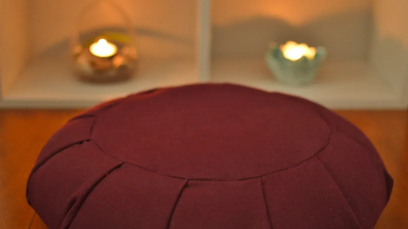 A maroon meditation cushion sits on a wooden floor, softly lit by two small, flickering candles in the background, creating a serene, tranquil atmosphere.