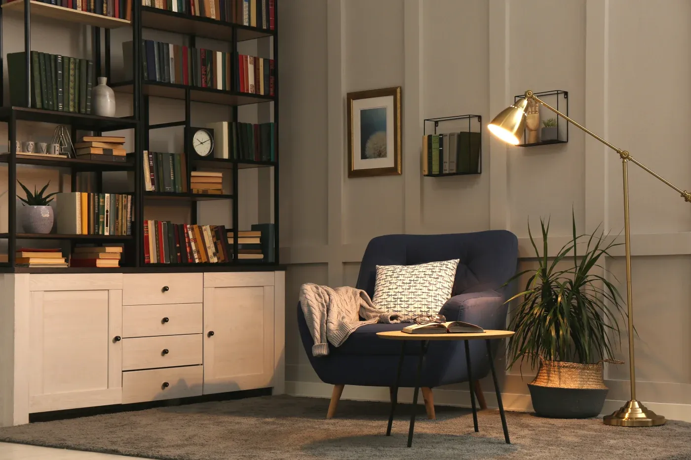Cozy reading nook with a blue armchair, patterned pillow, and draped blanket. A floor lamp shines over a small table with an open book, near a lush plant. Bookshelves filled with books and a clock line the wall, creating a warm, inviting atmosphere.