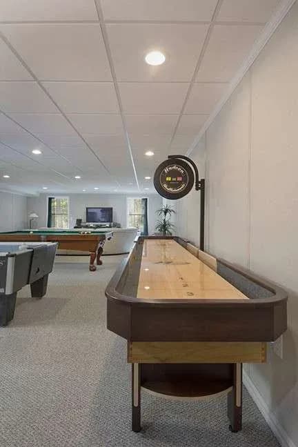 Basement game room with a shuffleboard table and a pool table. A TV and comfortable seating are visible in the background. The space is well-lit and inviting.
