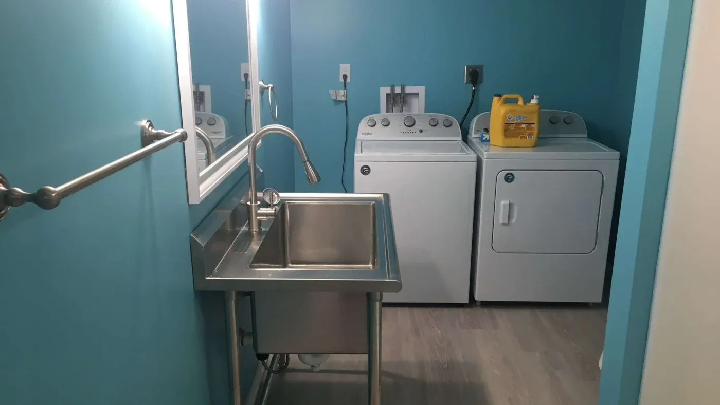A bright laundry room with light blue walls features a stainless steel sink on the left and a washer-dryer set on the right, with detergent on top.