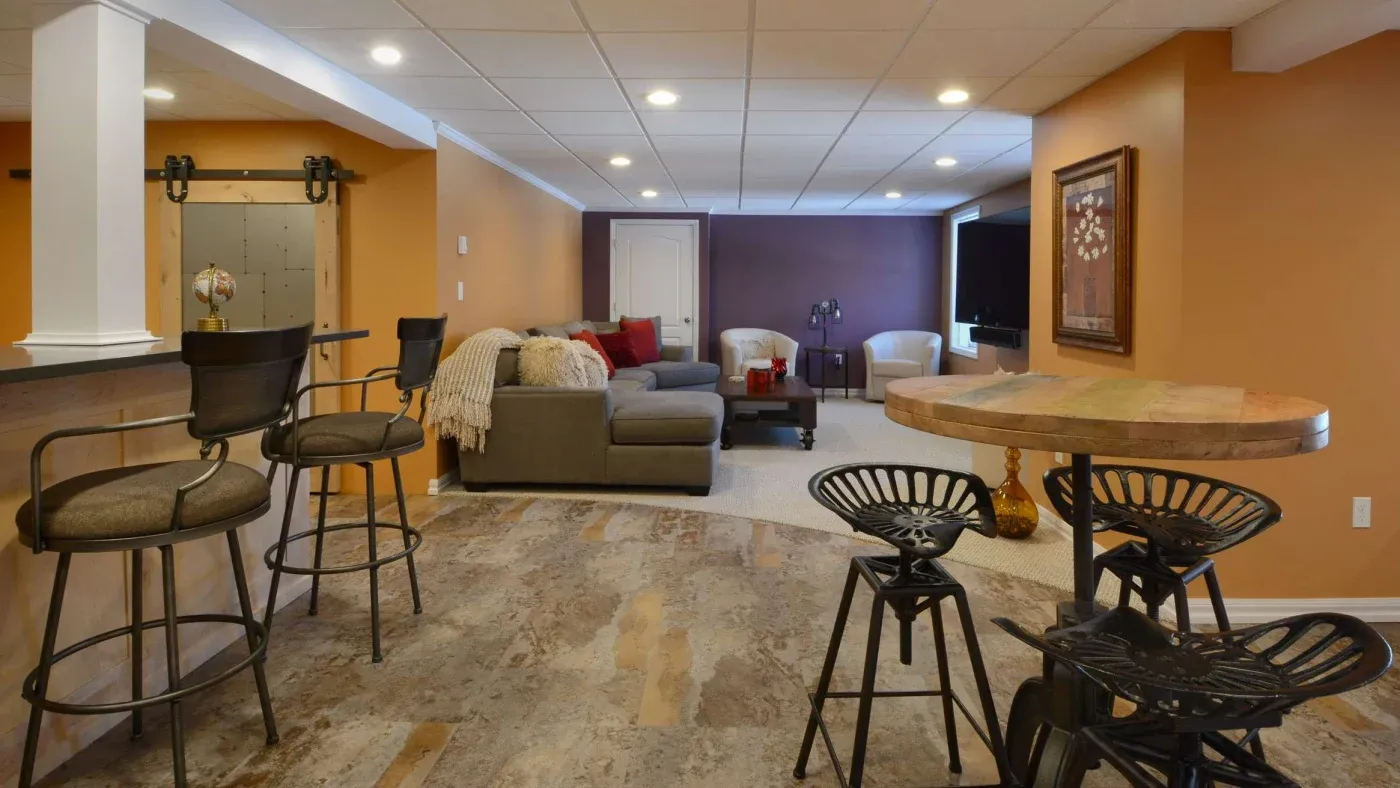 Spacious basement with warm tones, featuring a bar area with stools, a cozy gray sectional sofa, and modern decor including a round table and chairs.
