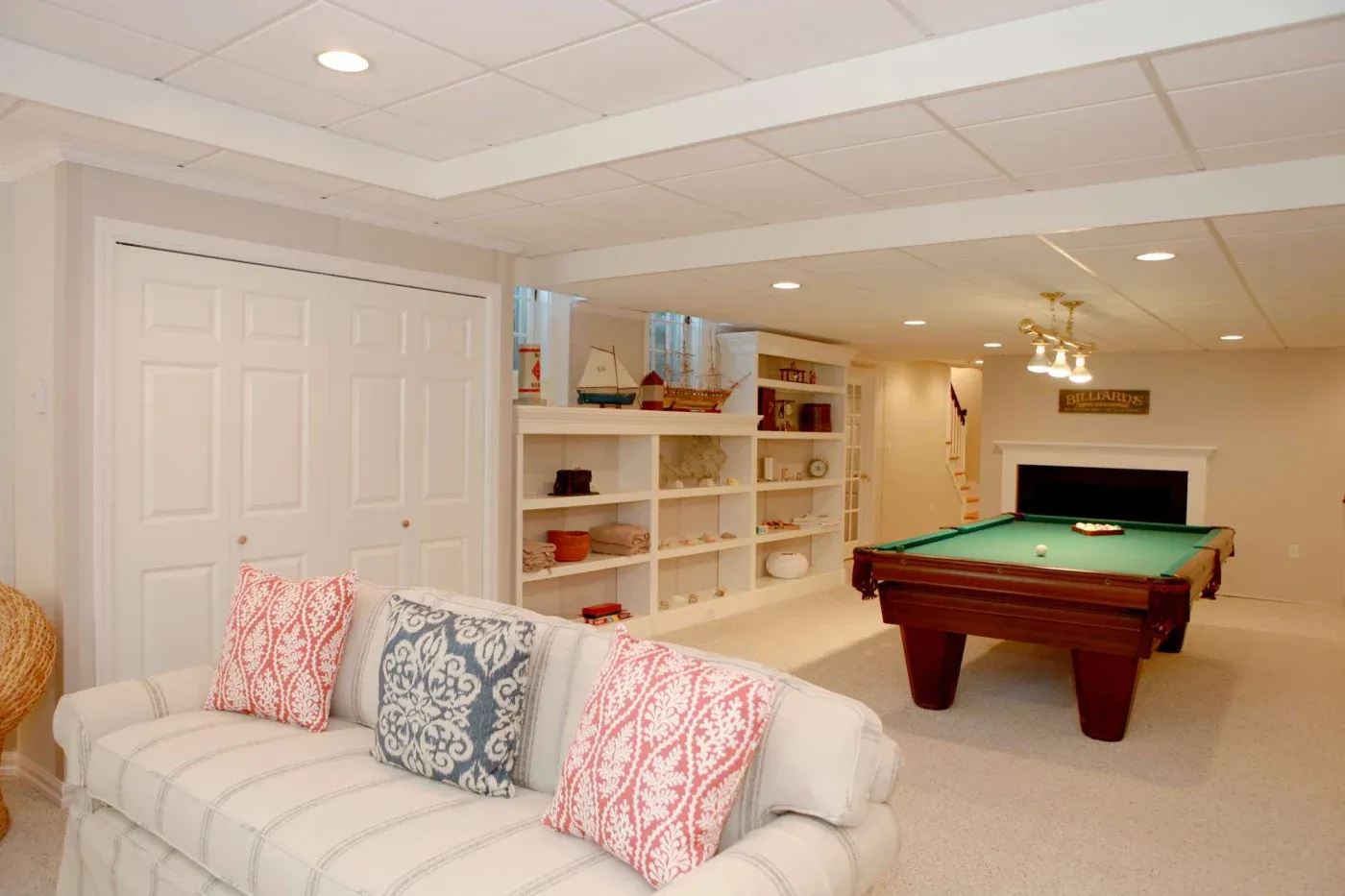 Cozy basement with a pool table under warm lighting. A beige sofa with red and blue patterned cushions faces bookshelves filled with decor items.