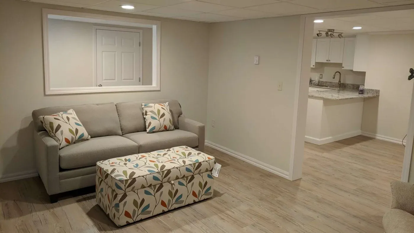 A cozy basement living area with light wood floors, a gray sofa with colorful leaf-patterned cushions, and a matching ottoman. A kitchen is seen in the background.