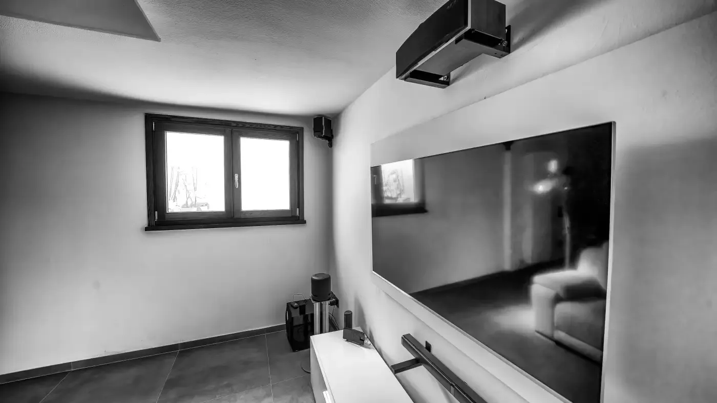 A modern room in grayscale featuring a large flat-screen TV on the wall. There's a window, minimal furniture, and a sleek, clean aesthetic.