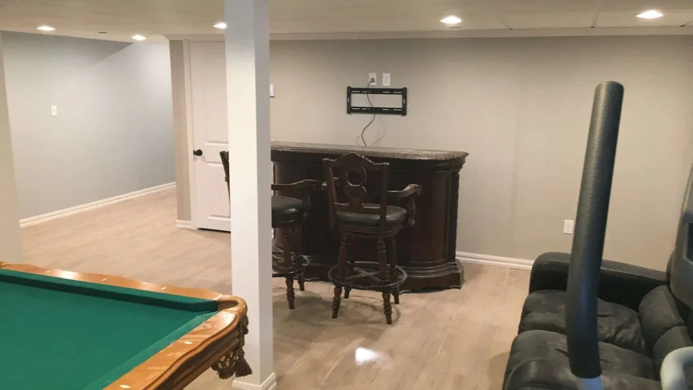 A modern basement features a green pool table, bar with ornate wooden chairs, and cozy black seating, conveying a relaxed, recreational atmosphere.