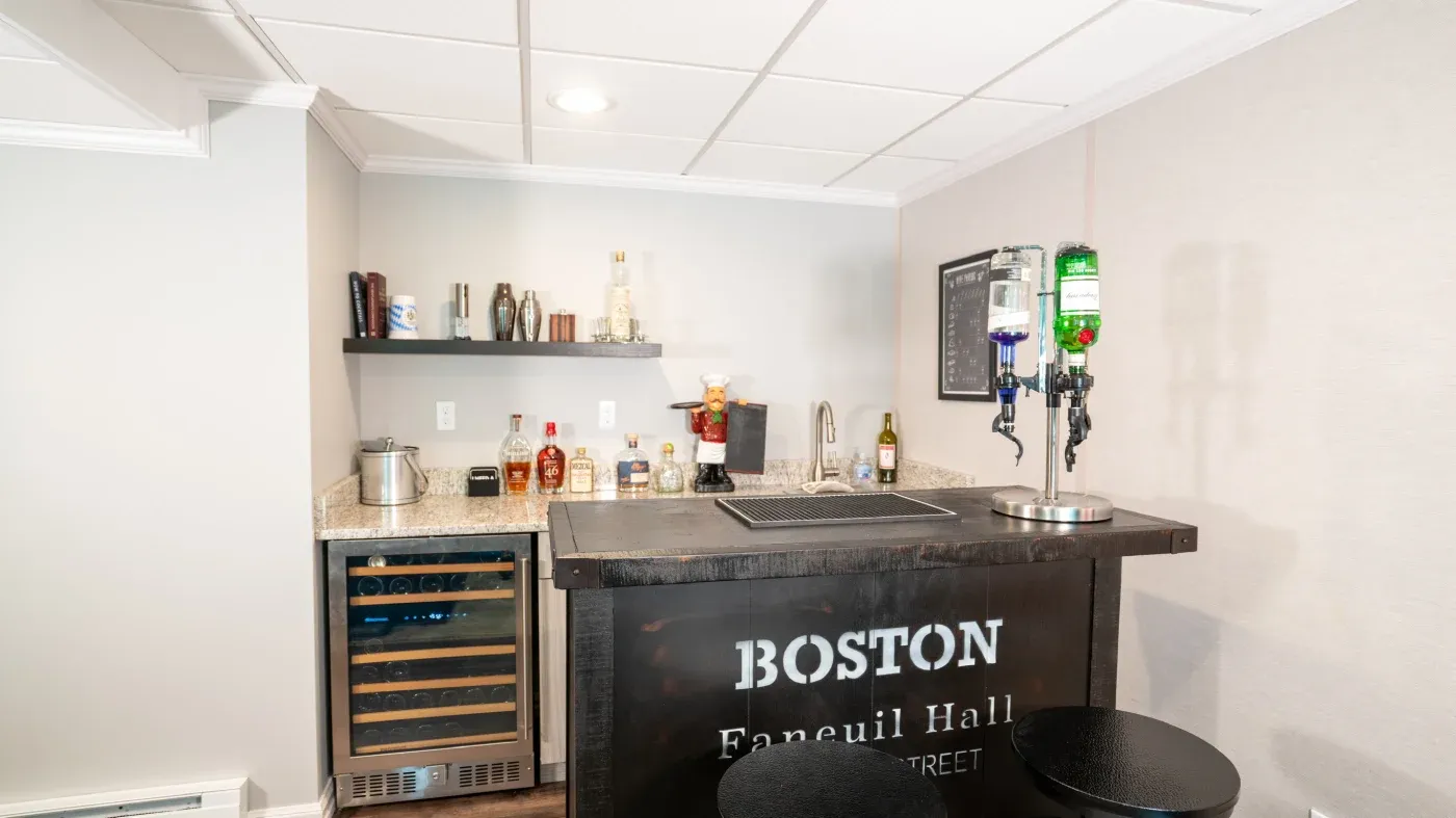 Modern home bar with "Boston Faneuil Hall" sign, featuring a black counter, liquor bottles on shelves, drink dispensers, and cozy ambiance.
