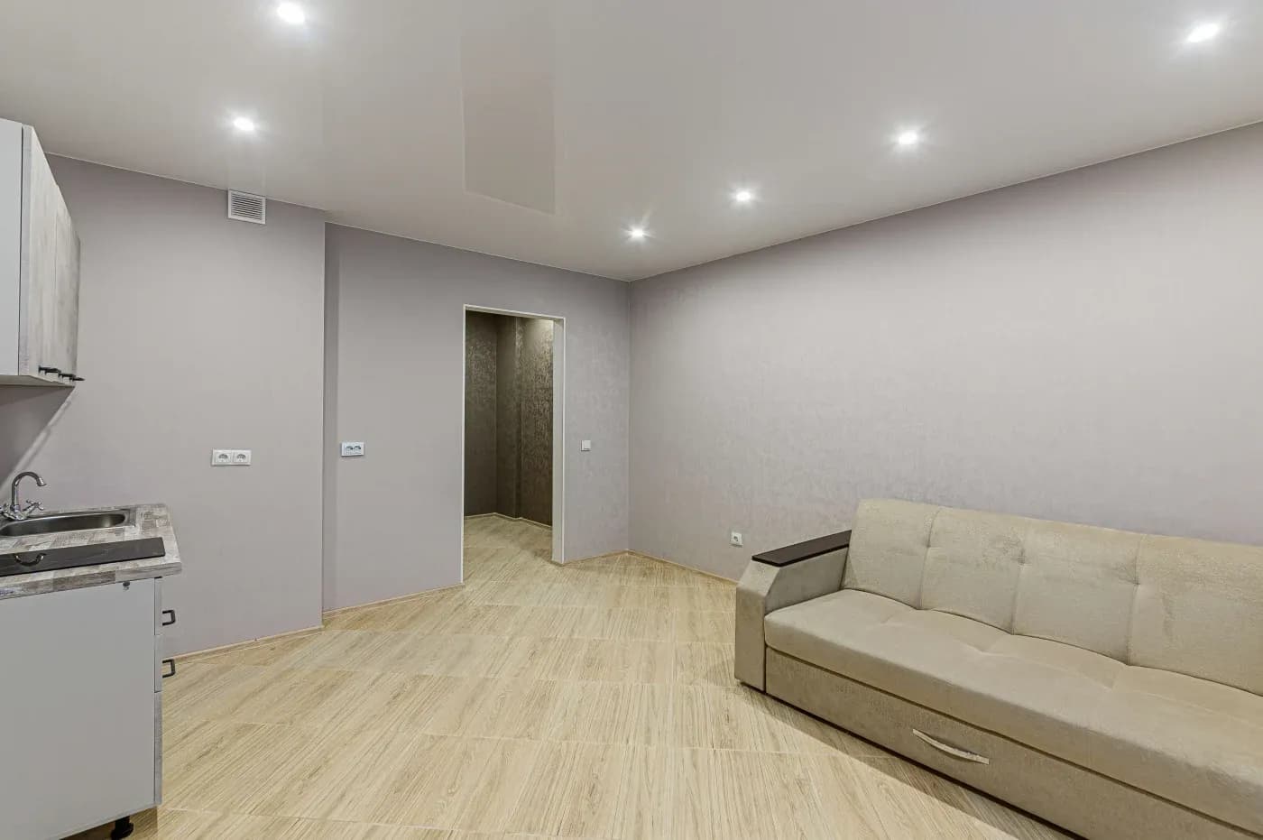 Minimalist room with light wood flooring, beige walls, and recessed lighting. Features a beige sofa and a small kitchenette with a sink.