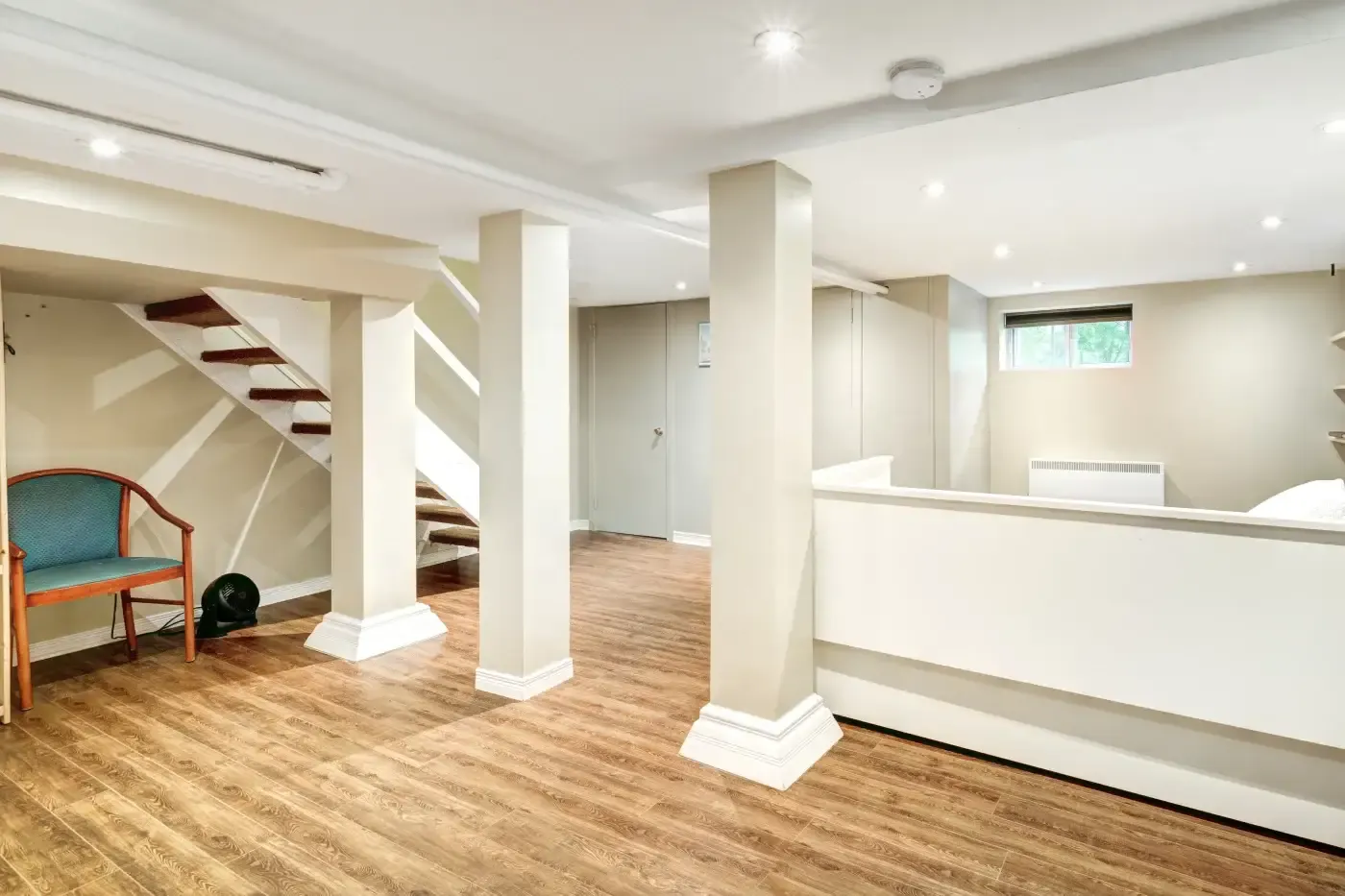 Basement with beige walls, wood flooring, and white columns. A wooden chair sits near stairs. A small window suggests natural light. Modern and tidy.