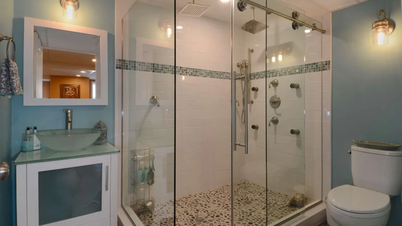 Modern bathroom with a large glass shower featuring multiple showerheads, pebble-style flooring, a turquoise vanity with a glass basin, and light blue walls.