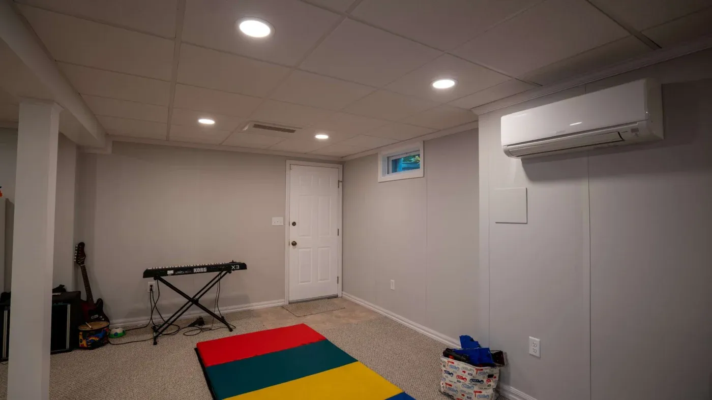 A minimalist basement with light gray walls, a colorful foam mat on the floor, a keyboard, and a guitar in the corner. The room feels calm and simple.