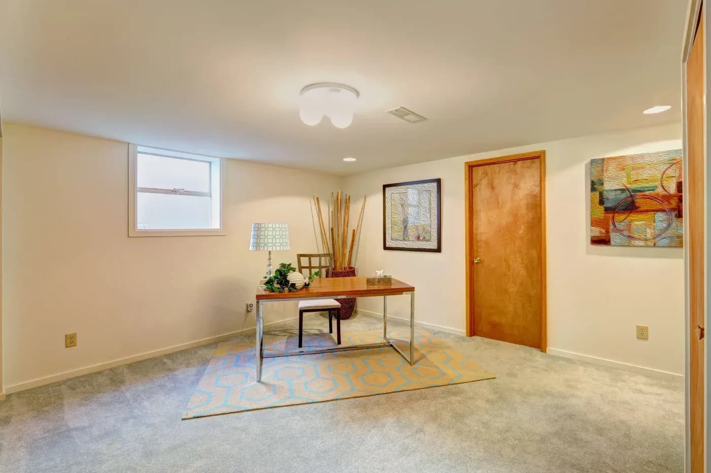 A minimalist beige room features a wooden desk with a plant and lamp, modern art on the walls, a patterned rug, and a closed wooden door. Calm and tidy ambiance.
