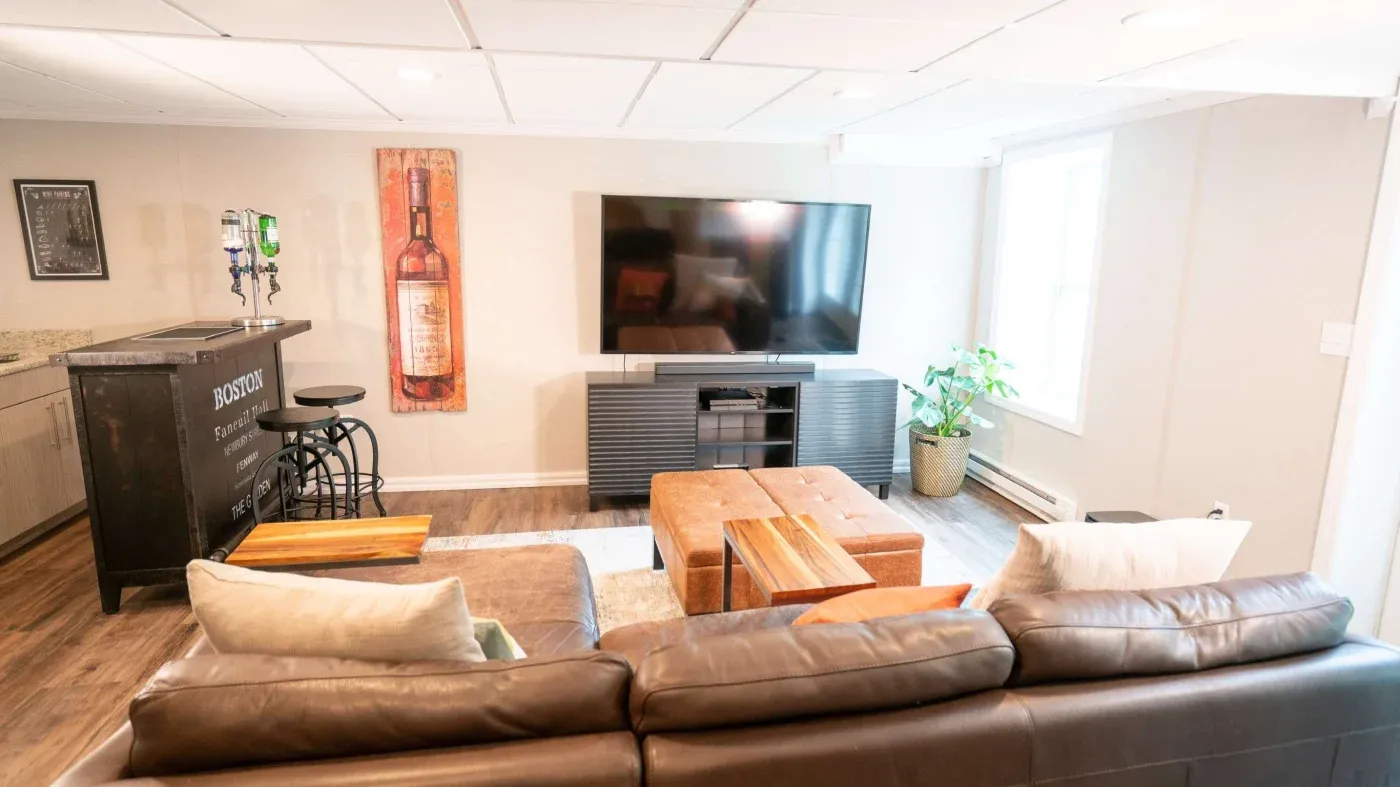 A cozy living room with a brown leather couch facing a large flat-screen TV. A small bar area with stools and bottle dispensers is on the left. A potted plant near the window adds a touch of greenery.