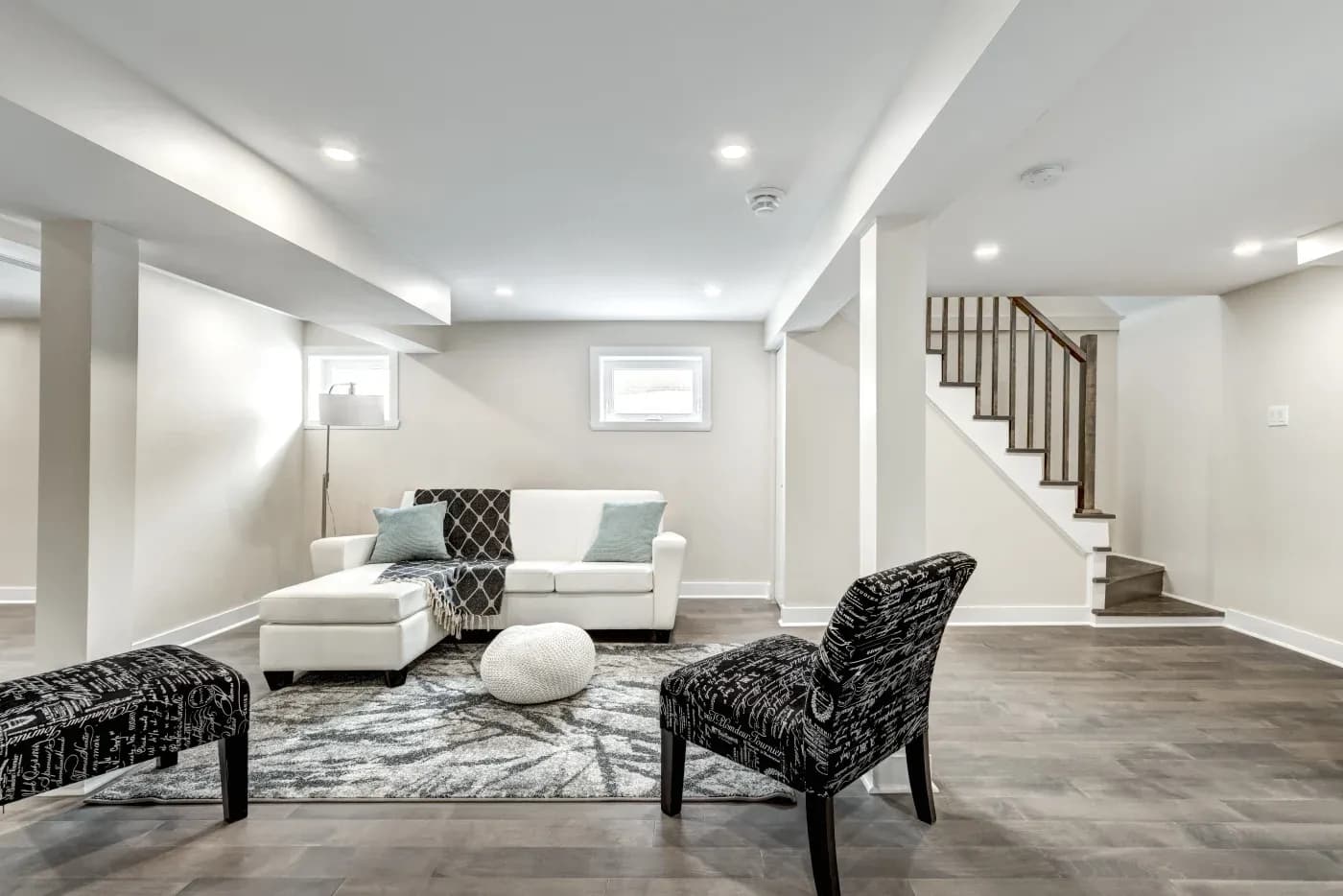 Modern basement features a white sofa with teal pillows, black patterned chairs, and a gray rug. Soft lighting and stairs add warmth to the sleek space.