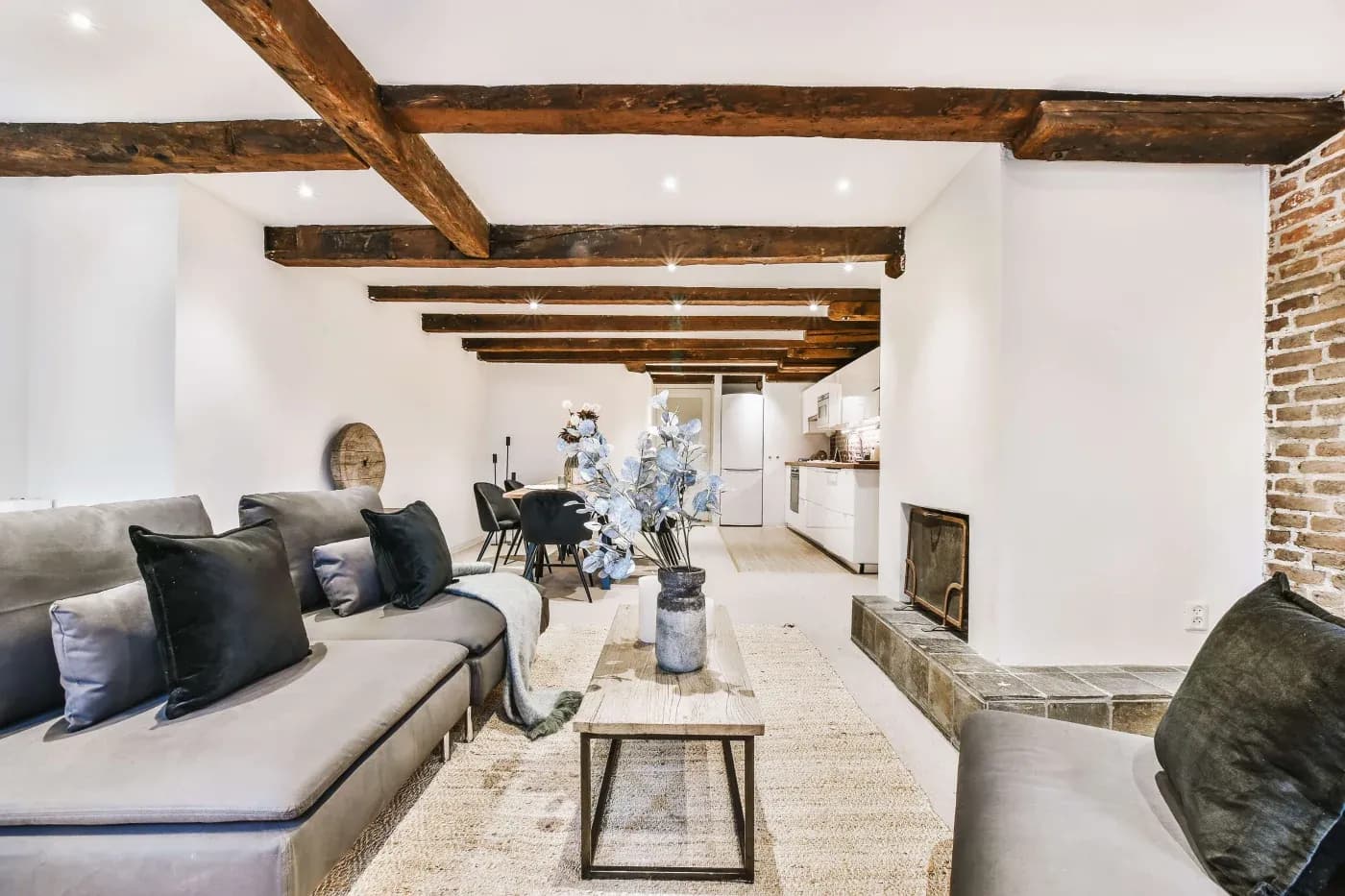 Modern living room with exposed wooden beams, white walls, and a brick accent. Grey sofas with black pillows surround a wooden coffee table with decorative branches. Open kitchen and dining area in the background. Cozy and stylish ambiance.