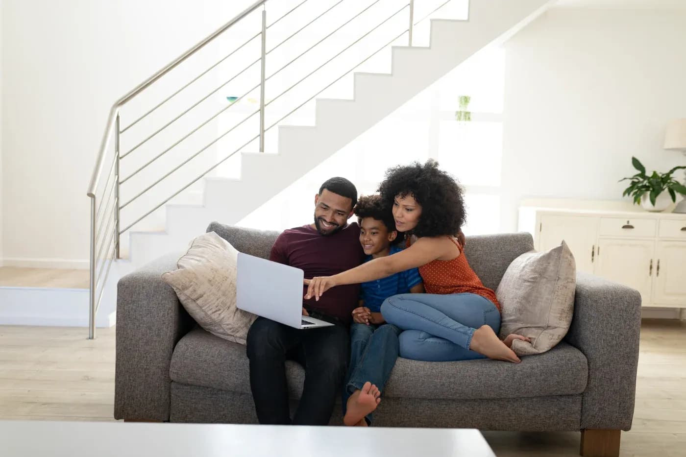A family of three sits on a gray sofa, smiling and looking at a laptop. The setting is a cozy living room with bright natural light and a staircase.