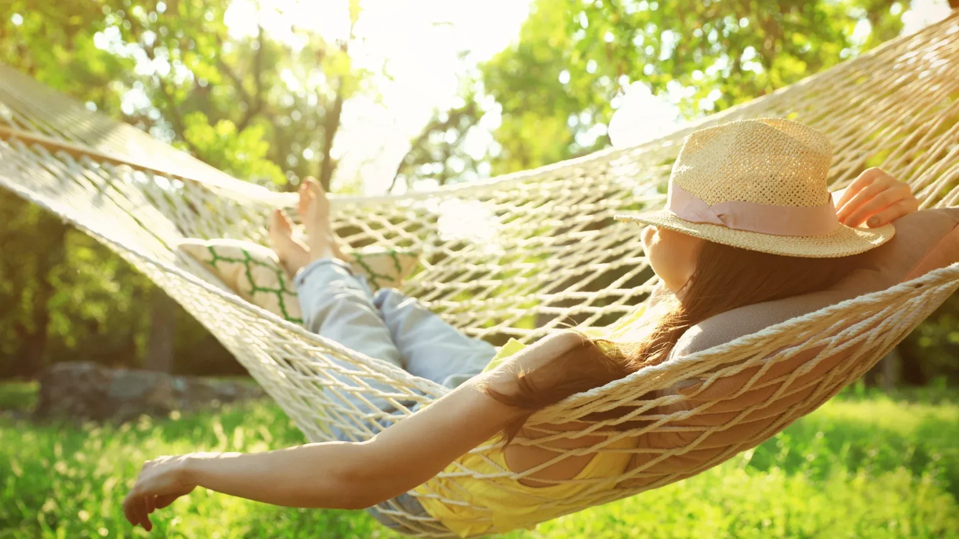Woman in a hat relaxing on a hammock in a sunlit garden, surrounded by lush green trees. The scene conveys tranquility and leisure.