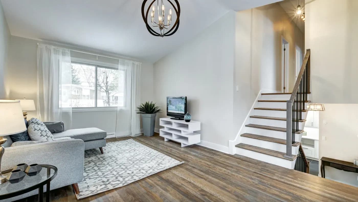 Modern living room with a gray sectional sofa, patterned rug, and flat-screen TV on a white stand. Wooden floors and a staircase enhance the cozy atmosphere.