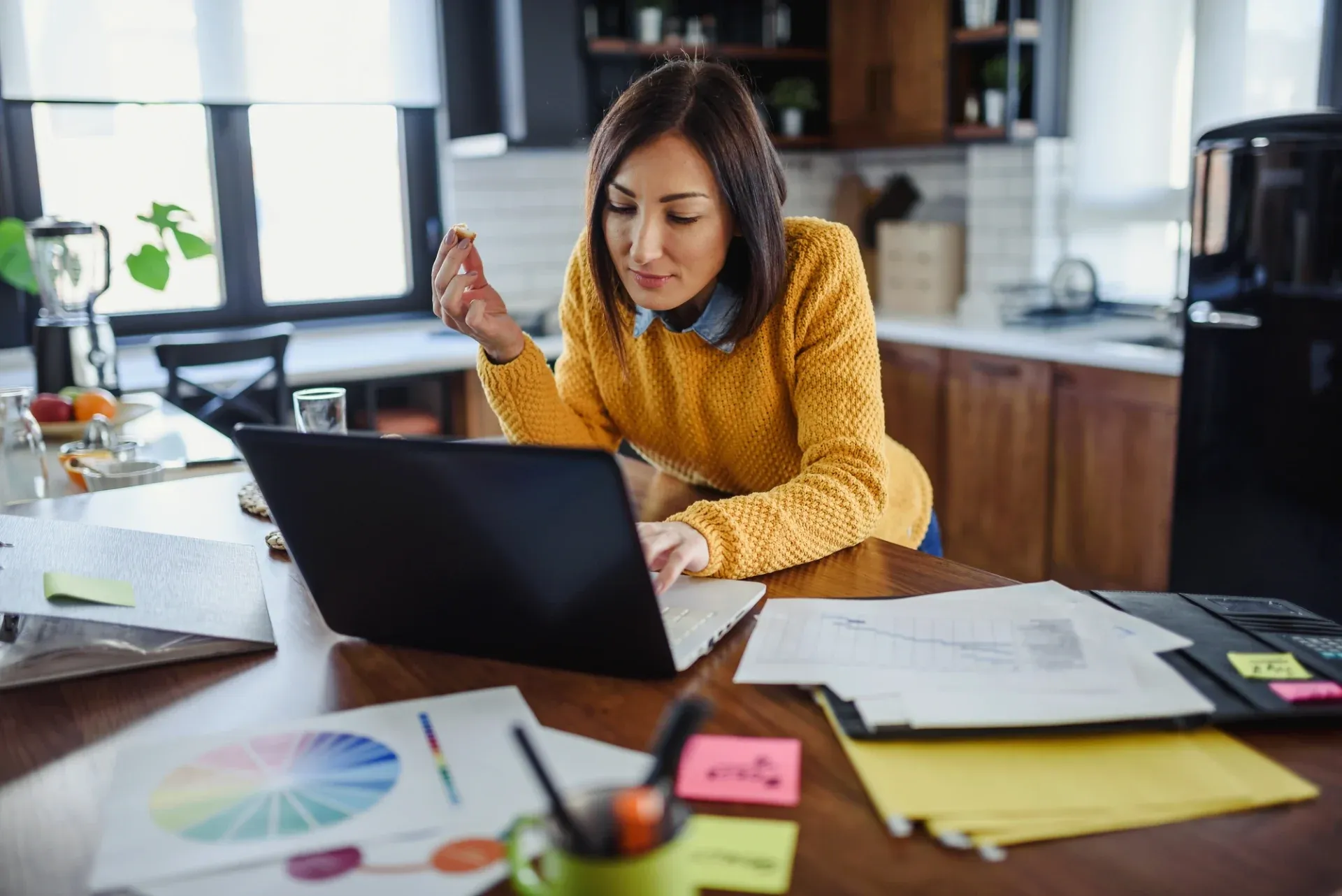 A woman in a yellow sweater intently works on a laptop in a cozy kitchen, surrounded by colorful charts and papers. The atmosphere is focused and productive.