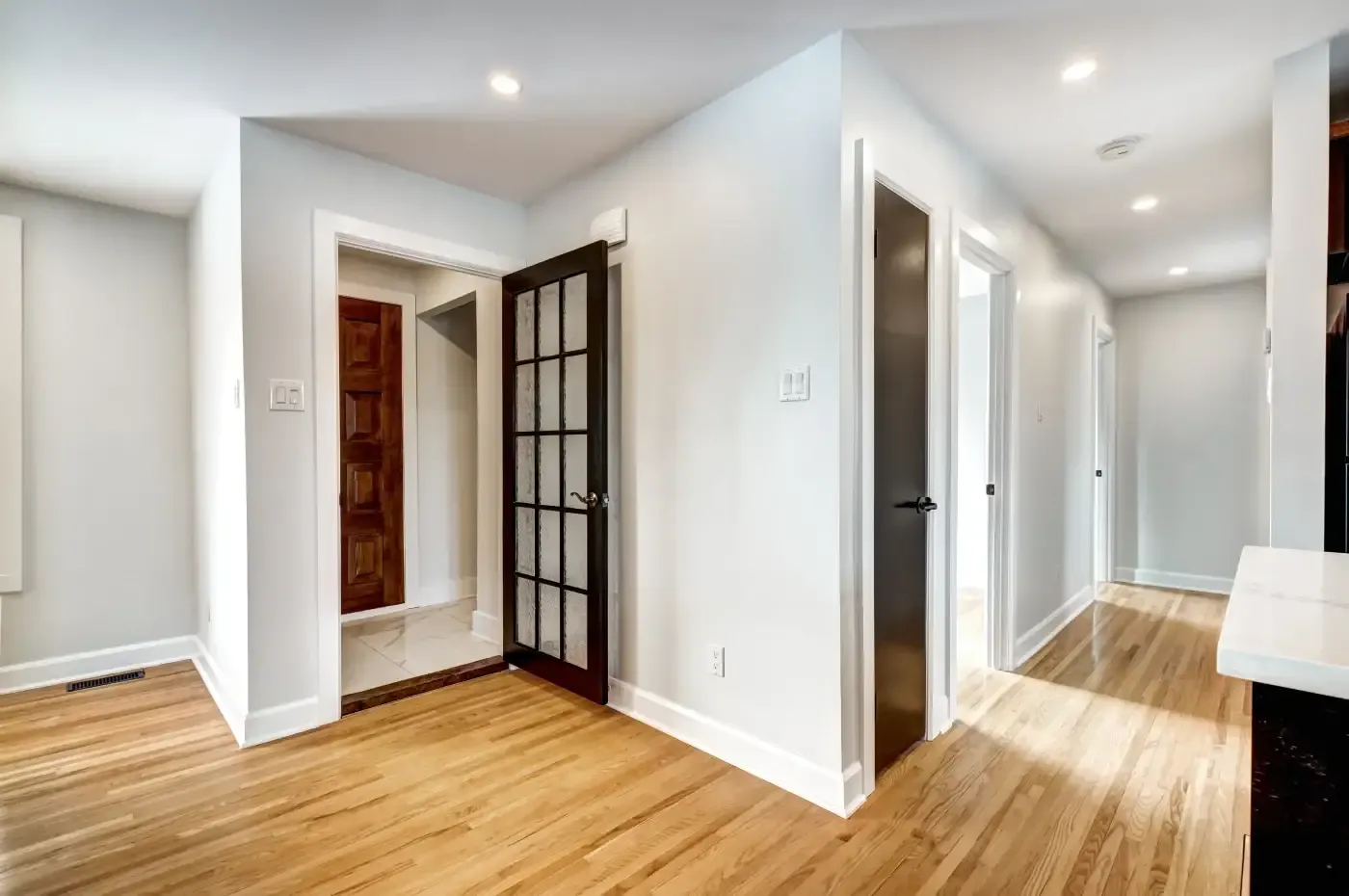 Bright hallway in a modern home with light hardwood floors, white walls, and recessed lighting. Features a glass-panel door and multiple doorways.