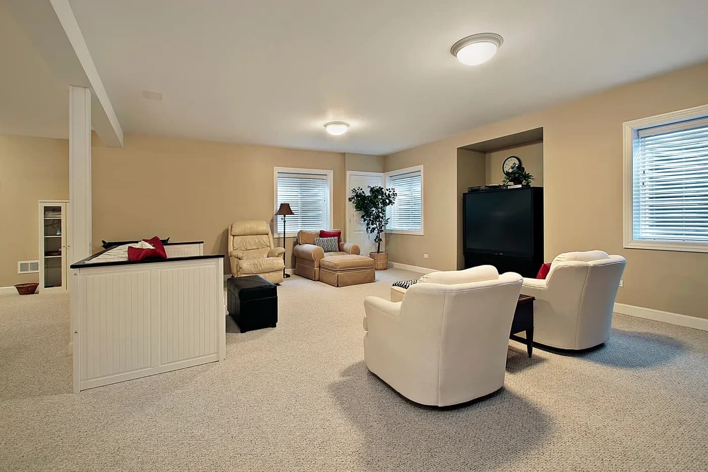 Spacious, beige living room with plush armchairs, a large TV, and a comfortable recliner. Soft lighting and a potted plant add warmth and coziness.