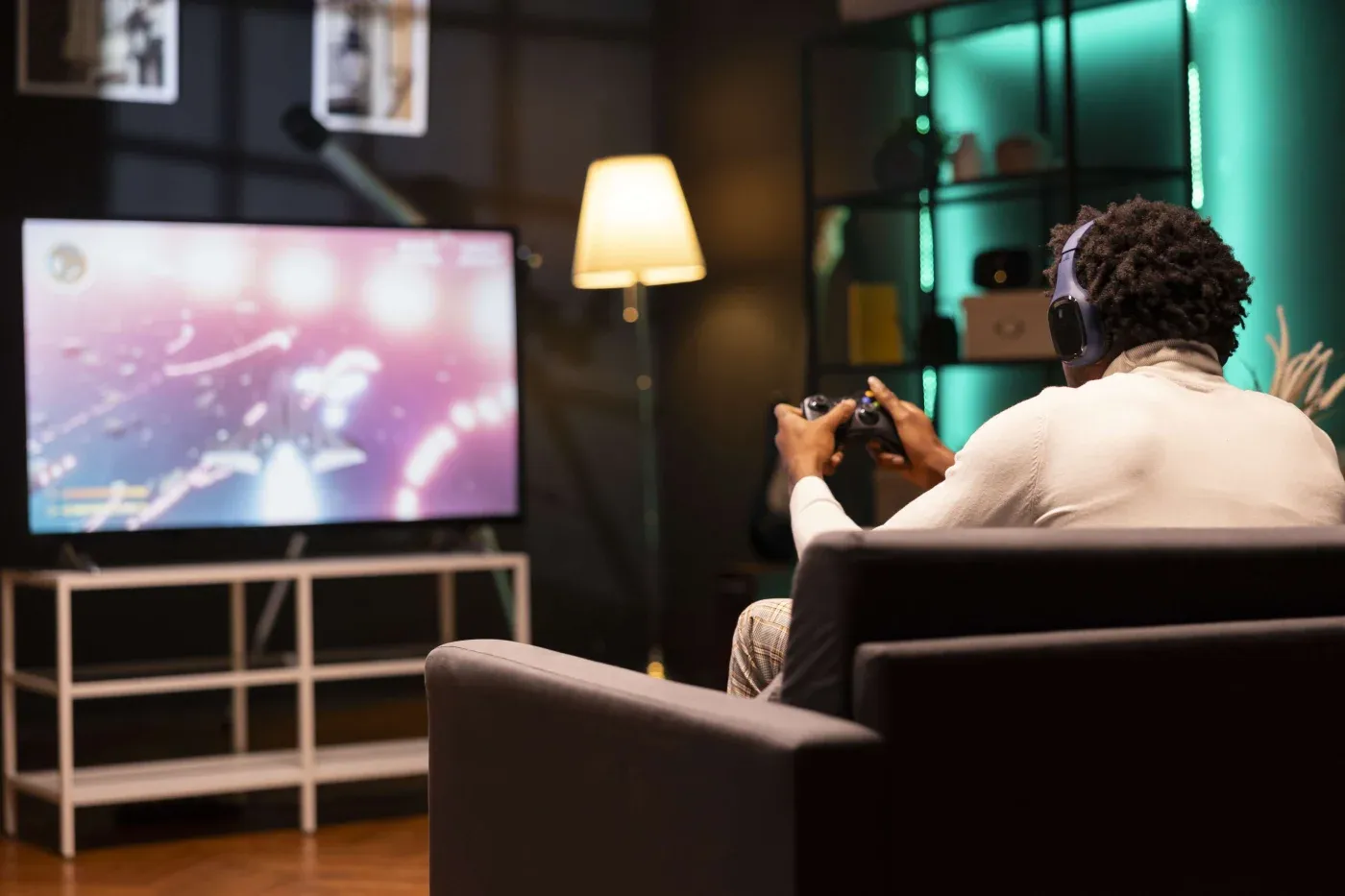 A person sits on a sofa in a dimly lit room, wearing headphones and playing a video game on a large TV. A soft lamp glows in the background.
