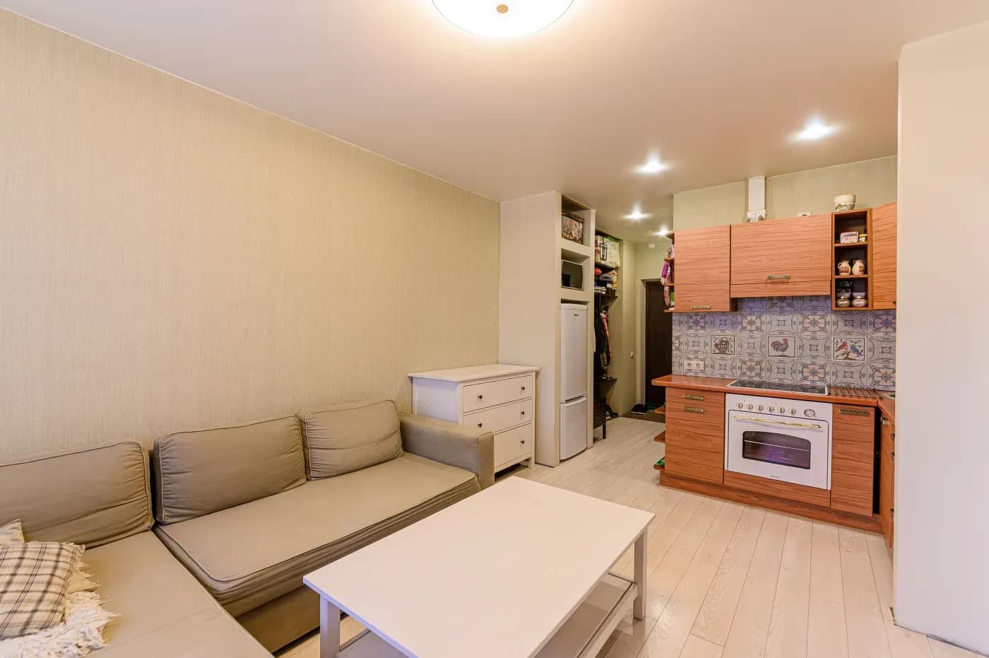 Compact living room with beige sofa, white coffee table, and integrated kitchen featuring wooden cabinets and patterned backsplash. Cozy, minimalist vibe.