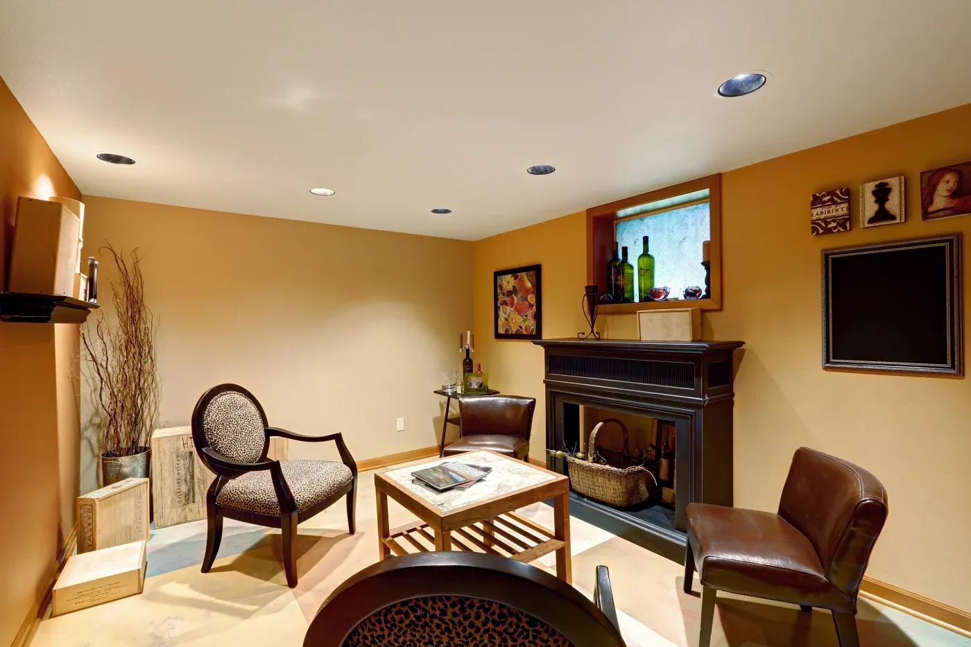 Cozy basement room with tan walls, featuring a fireplace, two leather chairs, a patterned armchair, a wooden coffee table, and decorative wall art.