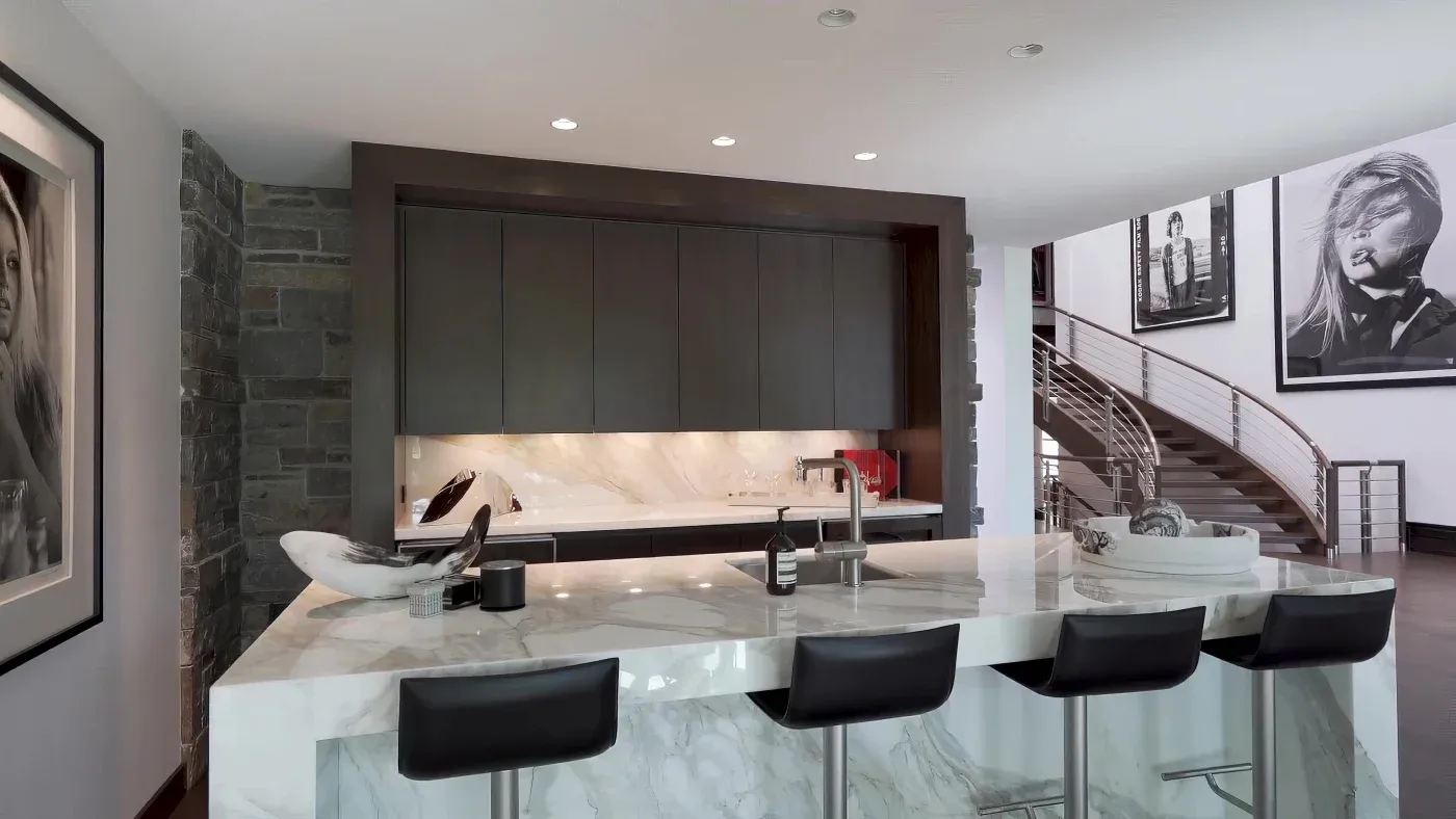 Modern kitchen with a large marble island and four black stools. Minimalist decor, sleek dark cabinets, and a spiral staircase in the background.