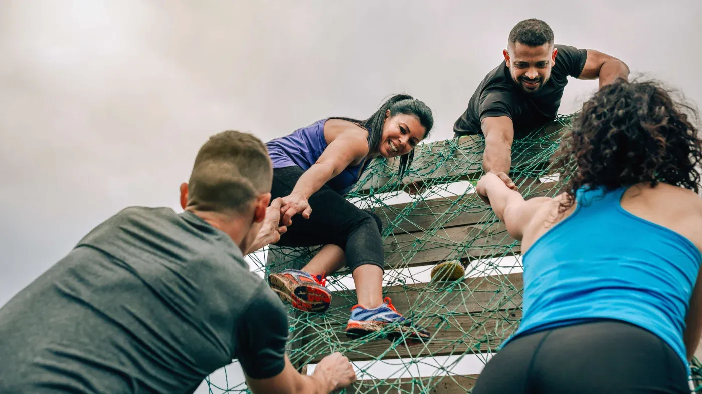 Four people participate in an outdoor obstacle course, helping each other climb a net wall. They exhibit teamwork and determination under a cloudy sky.
