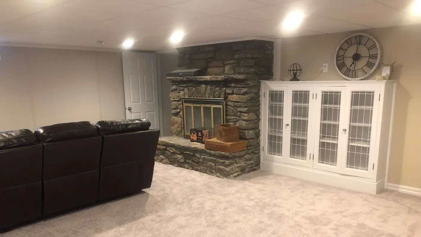 A cozy basement features a stone fireplace with logs and decor, a brown leather sofa, beige carpet, and a white cabinet with a large wall clock.