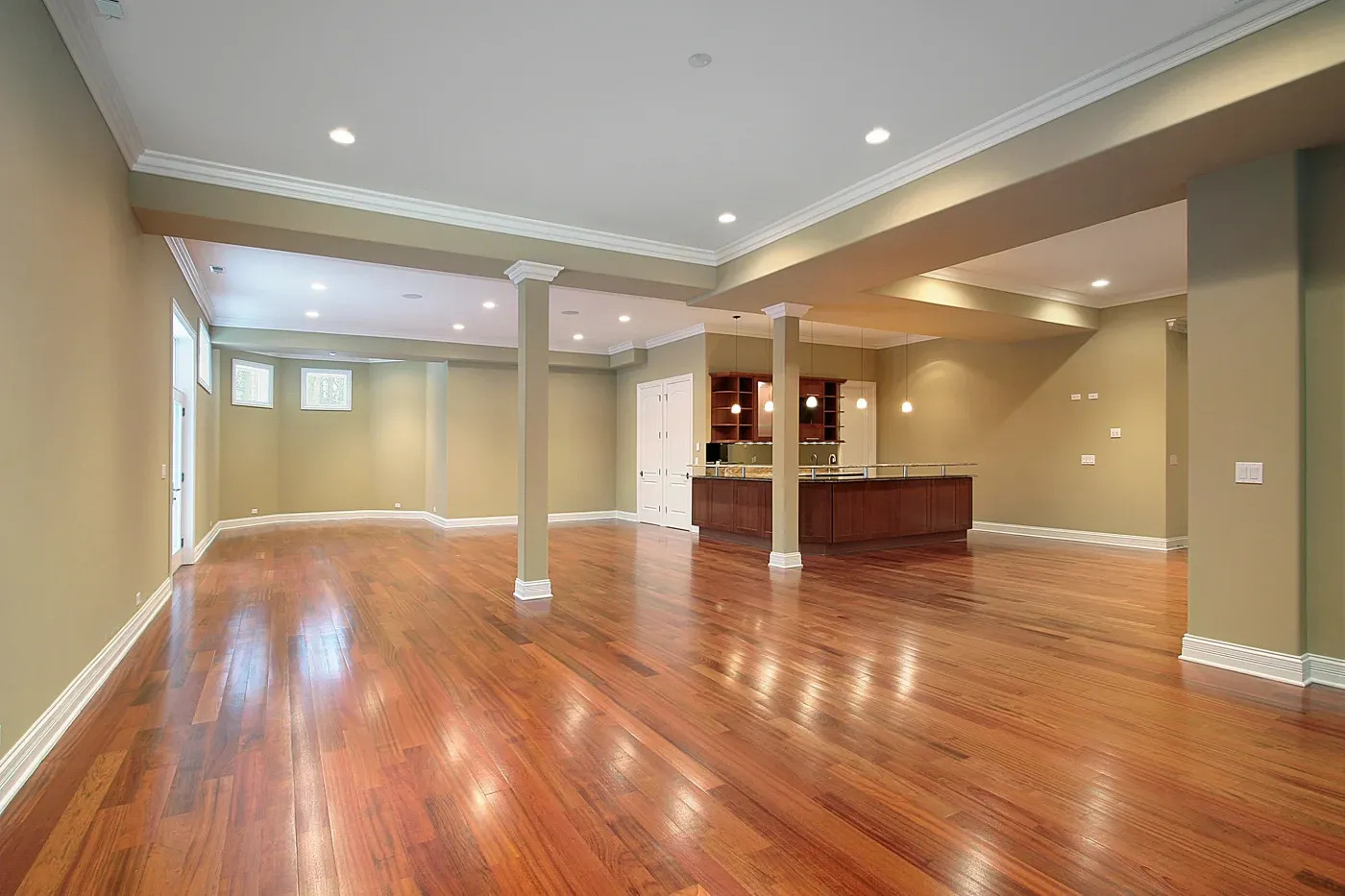 Spacious basement with polished wooden floors, tan walls, and recessed lighting. Features a small bar area with cabinets, creating a warm, inviting ambiance.