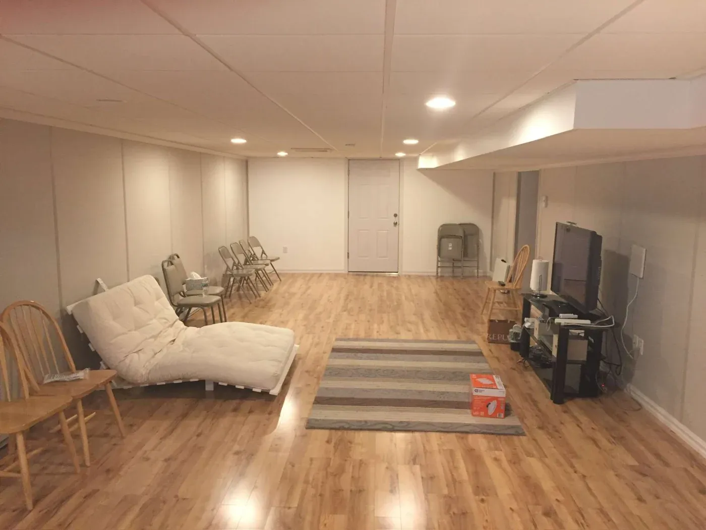 Basement with wood flooring, white walls, and overhead lighting. Features a TV, chairs, a reclined futon, and a striped rug, creating a simple, cozy atmosphere.
