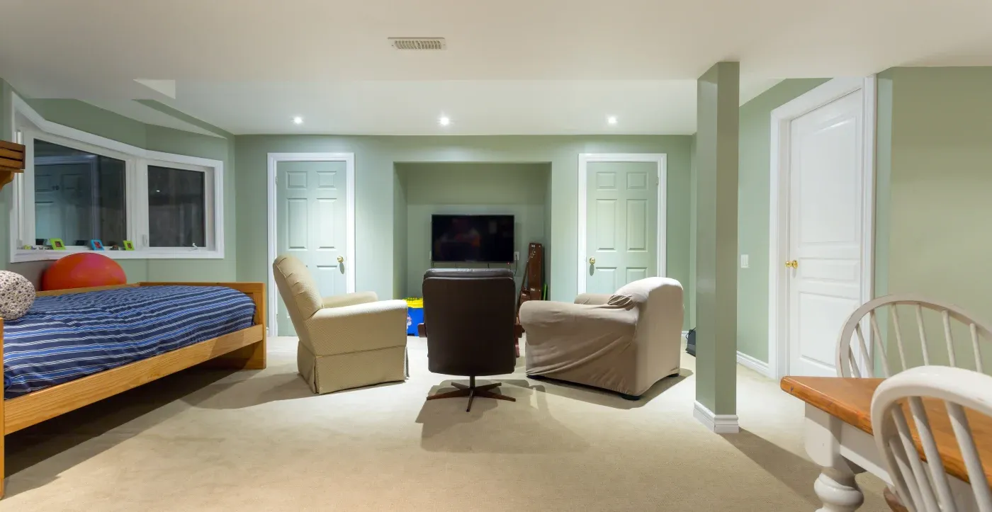 A cozy basement living area with a bed, armchairs, and a TV. Soft lighting and soothing green walls create a calm, inviting atmosphere.